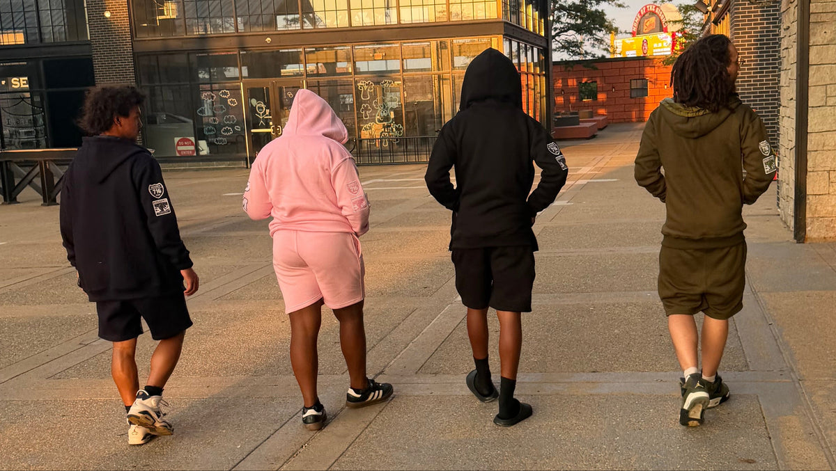 Four people walking away on a paved outdoor walkway at sunset, wearing matching hooded sweatshirt and shorts sets in different colors: black, light pink, black, and olive green.